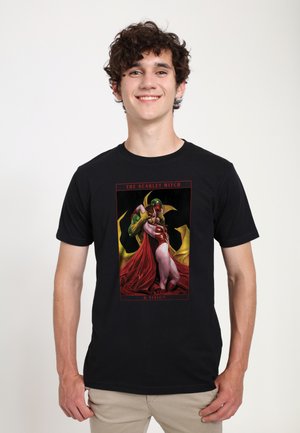Jeune homme portant un t-shirt noir avec un graphisme coloré de Scarlet Witch et Vision s'embrassant, debout devant un fond uni.