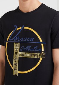 Svart bomullst-shirt med ett cirkulärt mönster med gula accenter och blå text som säger "Versace Collection" tillsammans med geometriska mönster.