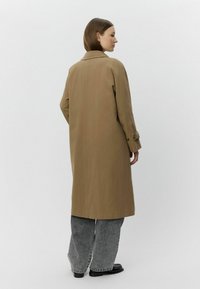 Barna hosszú kabát klasszikus gallérral, oldalsó gombokkal és egyenes szabással, laza szürke nadrággal és fekete cipővel párosítva.