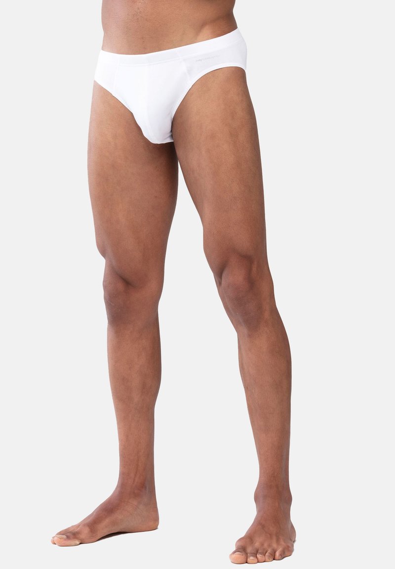 Slips en coton blanc avec une texture lisse, une taille élastique et une coupe classique, présentés sur un individu debout pieds nus.