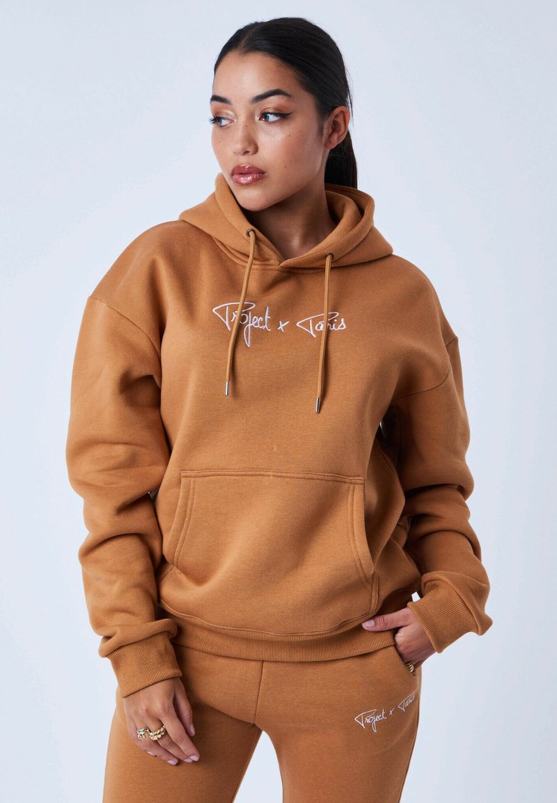 Bruine hoodie gemaakt van zachte stof, met een voorzak en witte logo borduursel. Ontspannen pasvorm met cuffs aan de mouwen.
