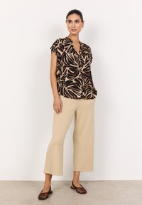 Eine Frau trägt eine kurzärmelige Bluse mit Blättermuster, beigefarbene weite Hosen und braune geflochtene Ballerinas und steht vor einer weißen Wand.