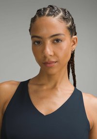 Zwarte mouwloze top met een V-halsontwerp. Het model heeft gevlochten haar en oorbellen met ringen. Gladde textuur tegen een neutrale grijze achtergrond.