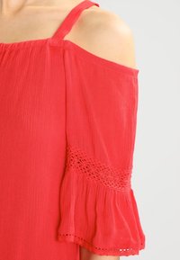 Haut rouge à épaules dénudées avec des bretelles fines, dotée de manches froncées et d'accents au crochet texturé le long de l'ourlet et des bords des manches.