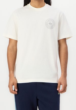Mann trägt ein schlichtes weißes Kurzarm-T-Shirt mit kleinem Lacoste Paris-Logo auf der linken Brust und dunkelblauer Hose.