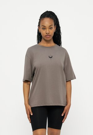 Donna con capelli intrecciati che indossa una maglietta grigia larga con una grafica di luna e stelle e pantaloncini da ciclista neri, in piedi davanti a uno sfondo semplice.