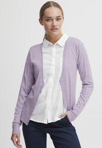 Cardigan violet clair en tissu tricot doux, avec un col en V, une ouverture sur le devant et des détails de boutons, aux manches longues, porté sur une chemise blanche.