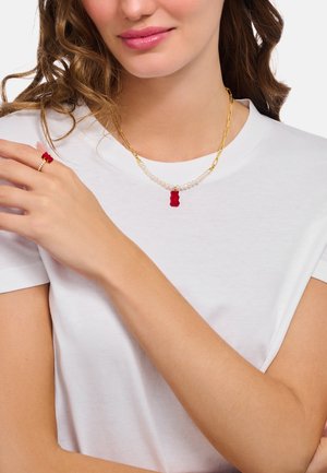 Frau im weißen Hemd, die eine goldene Halskette mit Perlen und einem roten Gummibärchen-Anhänger trägt, sowie einen passenden goldenen Ring mit rotem Gummibärchen.