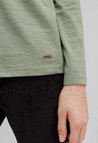 Camicia a maniche lunghe in cotone verde chiaro con orlo arrotondato e una piccola etichetta metallica. Abbinata a pantaloni in corduroy neri con texture.
