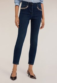 Jeans de mezclilla azul oscuro con una cintura alta, que presentan bolsillos decorados con detalles plateados y una silueta ajustada. Textura suave.