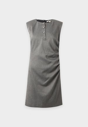 Frau trägt ein ärmelloses graues Kleid mit Knöpfen vorne, steht mit den Händen hinter dem Rücken und trägt hellfarbene High Heels.