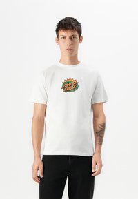 Witte katoenen t-shirt met een kleurrijk "Santa Cruz" grafisch ontwerp op de borst. Korte mouwen en ronde hals; model draagt zwarte jeans.