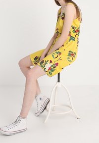 Gelbes Kleid mit Blumenmuster, dünnen Trägern und gerafftem Bund, kombiniert mit weißen High-Top-Sneakern, sitzend auf einem Hocker.