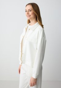 Witte zip-up jas van gladde stof met een kraag, gouden knopen, een voorzak en een geribde zoom, gecombineerd met bijpassende broek.