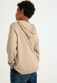 Beige hoodie, gemaakt van zacht materiaal, met een relaxte pasvorm, geribde zoom en manchet details; gedragen met donkerblauwe jeans, iets naar achter gekeerd.