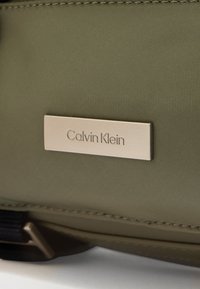 Τσάντα από νάιλον σε ελιά πράσινο χρώμα με ασημένια μεταλλική ετικέτα που φέρει χαραγμένο το "Calvin Klein". Λεία υφή με ραμμένες άκρες και ελάχιστα μεταλλικά στοιχεία.