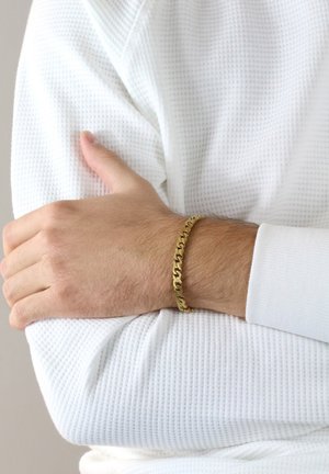 Gold chain bracelet on a wrist, featuring interconnected links. The bracelet has a smooth finish, worn over a textured white fabric.