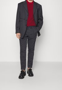 Grauer maßgeschneiderter Blazer über einem roten Strickpullover, kombiniert mit grauen Hosen mit Kordelzug und schwarzen Plateauschuhen. Glatter Stoff, funktionale Designdetails.