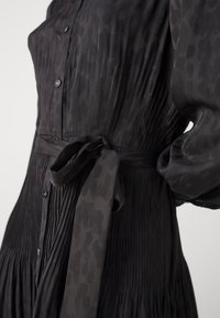 Vestido negro, plisado, con un sutil estampado de leopardo, cierre de botones y cinturón en la cintura. Las mangas largas y fluidas, junto con la textura sedosa, realzan el diseño.