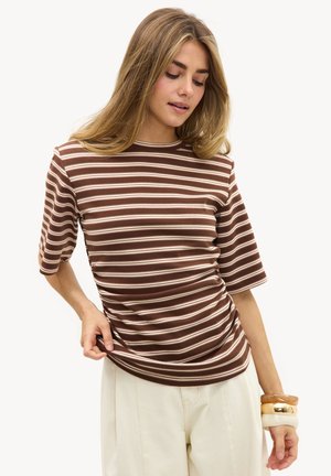 Femme portant un haut à manches courtes rayé marron et blanc, un pantalon beige, et plusieurs bracelets épais, regardant vers le bas en ajustant l'ourlet de sa chemise avec la main.