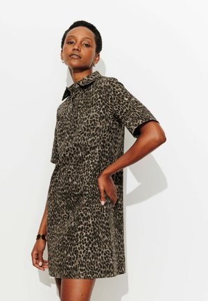 Femme posant devant un fond blanc portant une robe à manches courtes avec col, imprimé léopard, main sur la hanche.