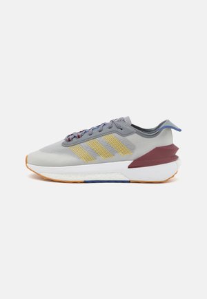 Zapatilla deportiva de color gris claro con rayas amarillas, acentos en el talón color marrón, parte superior de malla texturizada y una suela blanca acolchada con detalles en naranja y azul.