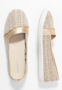 Chaussures slip-on tissées beige avec une bande métallique dorée sur le dessus et des semelles en caoutchouc blanches, présentant un design de tissu texturé.