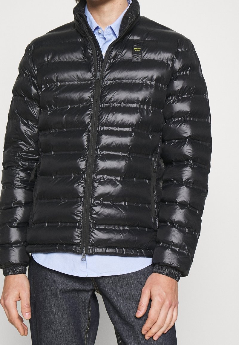 Giubbotto imbottito nero con superficie lucida e strutturata, trapuntatura orizzontale, colletto alto e chiusura frontale con zip. Materiale morbido e polsini aderenti.
