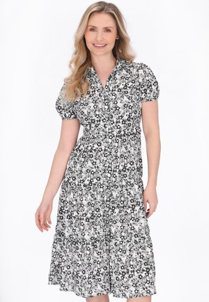 Kvinde med langt blondt hår iført en sort og hvid blomsterprintet midi-kjole med korte ærmer, smilende og stående mod en hvid baggrund.