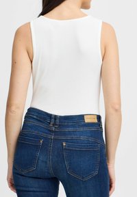 Bílý žebrovaný top bez rukávů v kombinaci s modrými džíny, které mají béžovou koženou nášivku a prošívané kapsy.