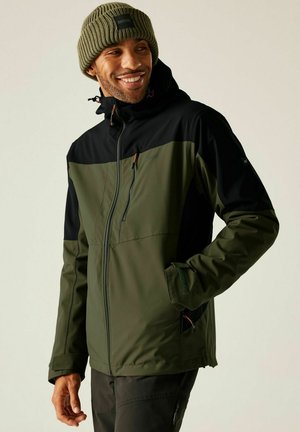 Mężczyzna uśmiechający się, ubrany w zielono-czarną outdoorową kurtkę na zamek i zieloną dzianą czapkę, ręce w kieszeniach kurtki, patrzący lekko na bok.