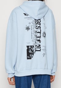 Jasnoniebieska, oversize bluza z kapturem z miękkiej tkaniny, zdobiona czarnymi nadrukami graficznymi, w tym wzorami oczu, słońca oraz elementami kwiatowymi.