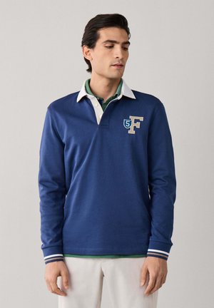 Homme portant un maillot de rugby bleu à manches longues avec un col blanc, un pantalon beige et une chemise verte en dessous, debout devant un fond uni.