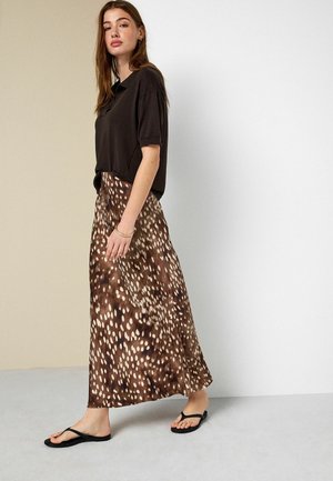 Woman walking wearing black loose short-sleeve polo shirt, brown and white spotted long skirt, and black flip-flops on gray floor.