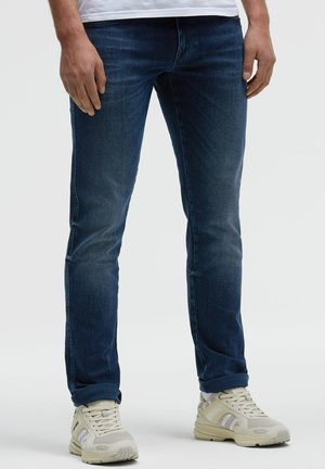 Mann trägt dunkelblaue Slim-Fit-Jeans und beige Sneaker und steht vor einfachem Hintergrund.