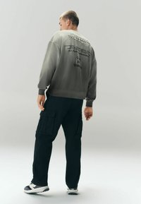 Homme aux cheveux courts vu de dos, portant un sweat-shirt gris avec du texte noir et un pantalon cargo noir avec des poches.
