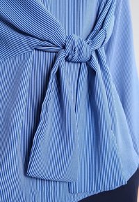 Modrá a bílá látka s vertikálními pruhy svázaná uzlem s dlouhými volnými konci, nošená jako součást oděvu.