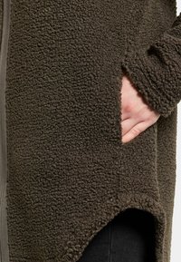 Mörkbrun fleecejacka med en strukturerad yta, som har en sidoficka och en asymmetrisk nederkant. Mjuk tyg och avslappnad passform.