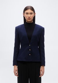 Μπλε ν αυτικό χειροποίητο σακάκι με βαθύ V-λαιμόκοψη, δομημένους ώμους και κλείσιμο με τέσσερα χρυσά κουμπιά. Υφή υφάσματος και εφαρμοστή σιλουέτα.