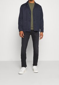 Marinblå skjorta med knappar över en olivgrön skjorta, i kombination med mörka jeans och vita sneakers. Tyget ser slätt ut och har en avslappnad design.