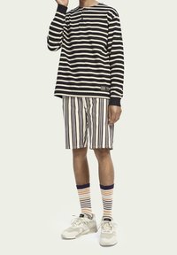 Schwarz-weiße gestreifte Langarmbluse, dazu passende gestreifte Shorts, farbenfrohe gestreifte Socken und helle Sneaker mit texturierter Oberfläche.