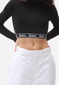 Musta pitkähihainen crop-top, jossa on valkoinen elastinen nauha, jossa lukee "Dickies"-logo, yhdistettynä korkeavyötäröisiin valkoisiin housuihin.