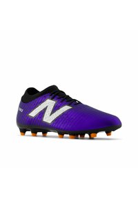 Botín de fútbol púrpura con una superficie texturizada, acentos en negro y un logotipo blanco sobredimensionado. Presenta tacos naranjas para tracción en el césped.