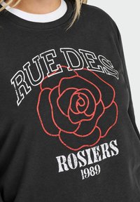 Sudadera negra con un gran contorno de rosa roja y el texto blanco "RUE DES ROSIERS 1989" en el pecho.