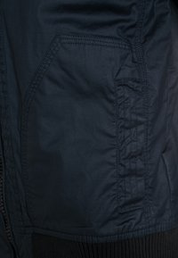 Close-up of a dark blue jacket pocket with detailed stitching and a black elastic cuff at the bottom edge.