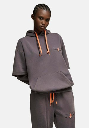 Jeune personne portant un sweat à capuche gris foncé à manches courtes et un pantalon assorti avec des cordons orange et noirs, debout devant un fond uni.