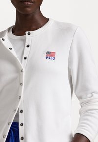 Detailní záběr na osobu v bílé košili na knoflíky, na které je vyšitá malá americká vlajka a nápis "POLO" na hrudi.