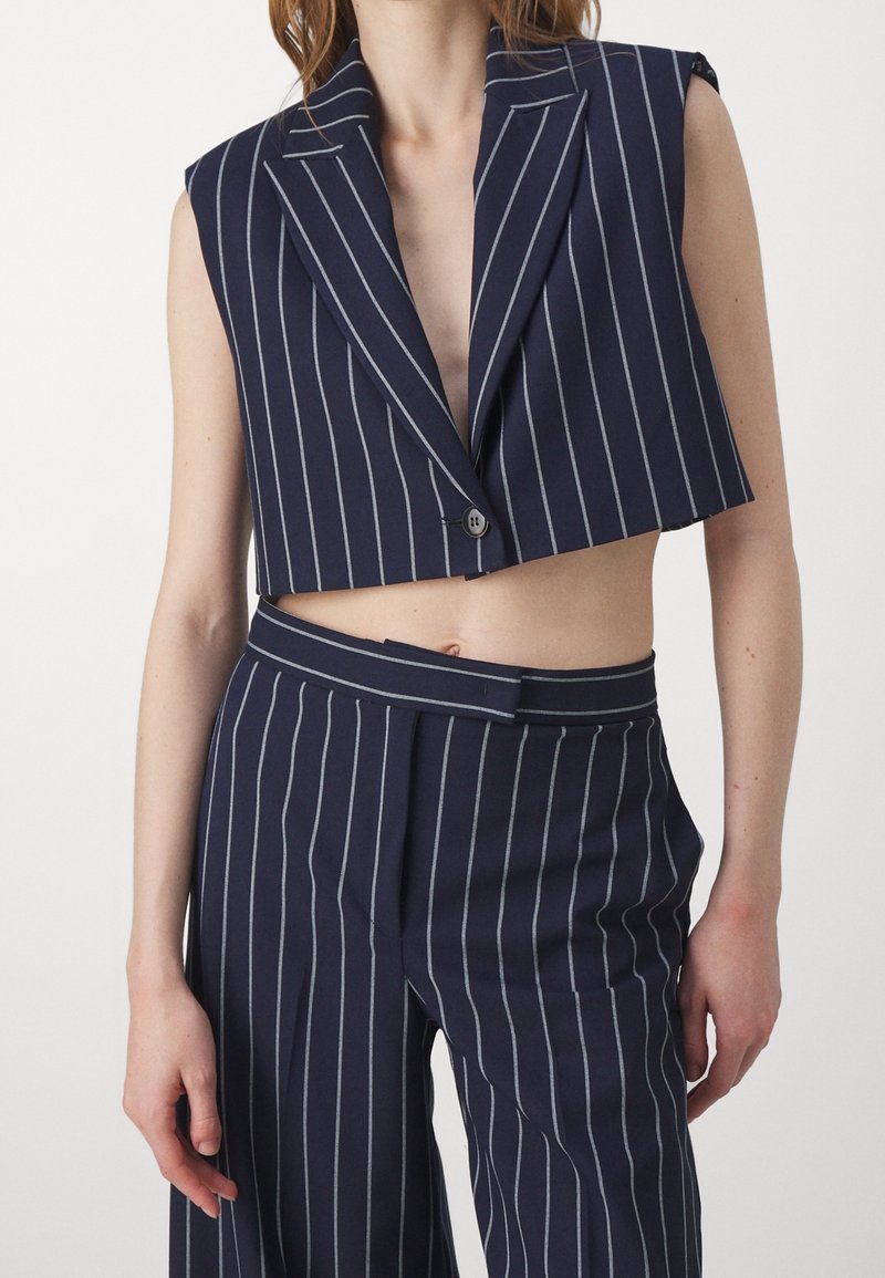 Marinblå kort blazer med vertikala vita ränder, skarpa slag och en enda knapp. Matchas med högmidjade randiga byxor.