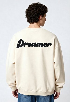 Man met krullend haar, die een crèmekleurige trui draagt met zwarte "Dreamer" tekst op de achterkant, gezien van achteren tegen een effen achtergrond.