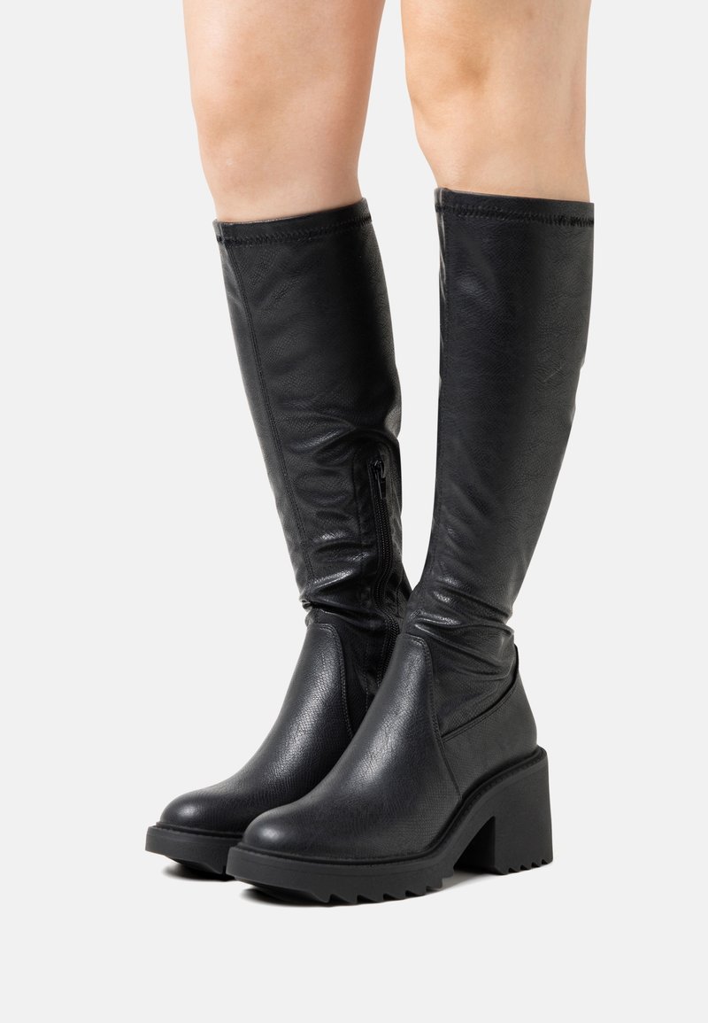 Bottes en cuir noires montant jusqu'au genou avec talons épais et fermetures éclair latérales portées sur des jambes nues, sur un fond uni.